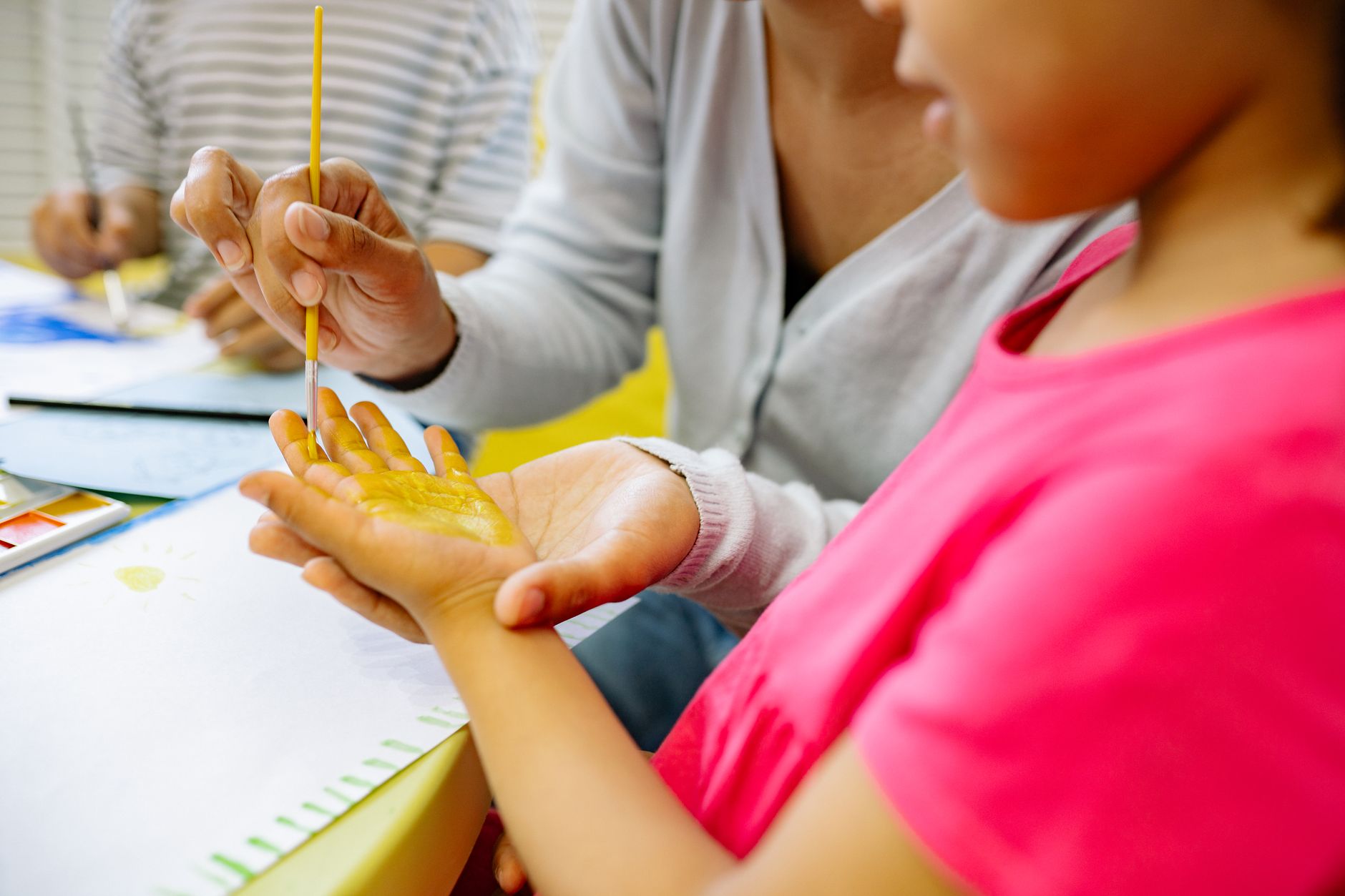 A teacher and young child painting the child's hand with yellow watercolor at a classroom table