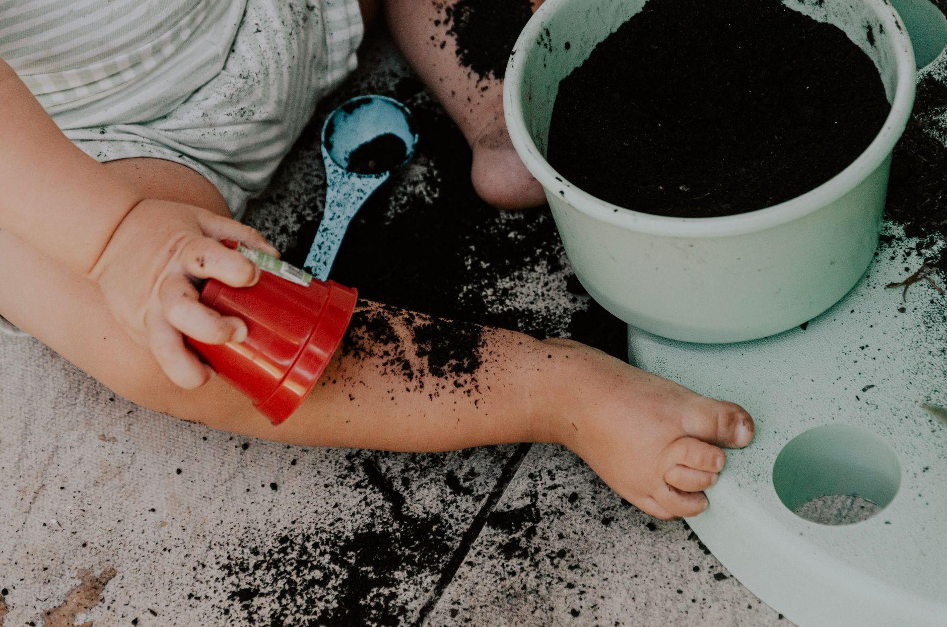 A toddler sitting on the ground exploring soil with a bucket and cup